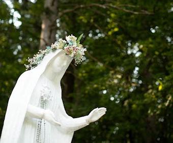 Our Lady of Fatima Statue adorned with beads and a flower crown with a backdrop of green tree foliage.